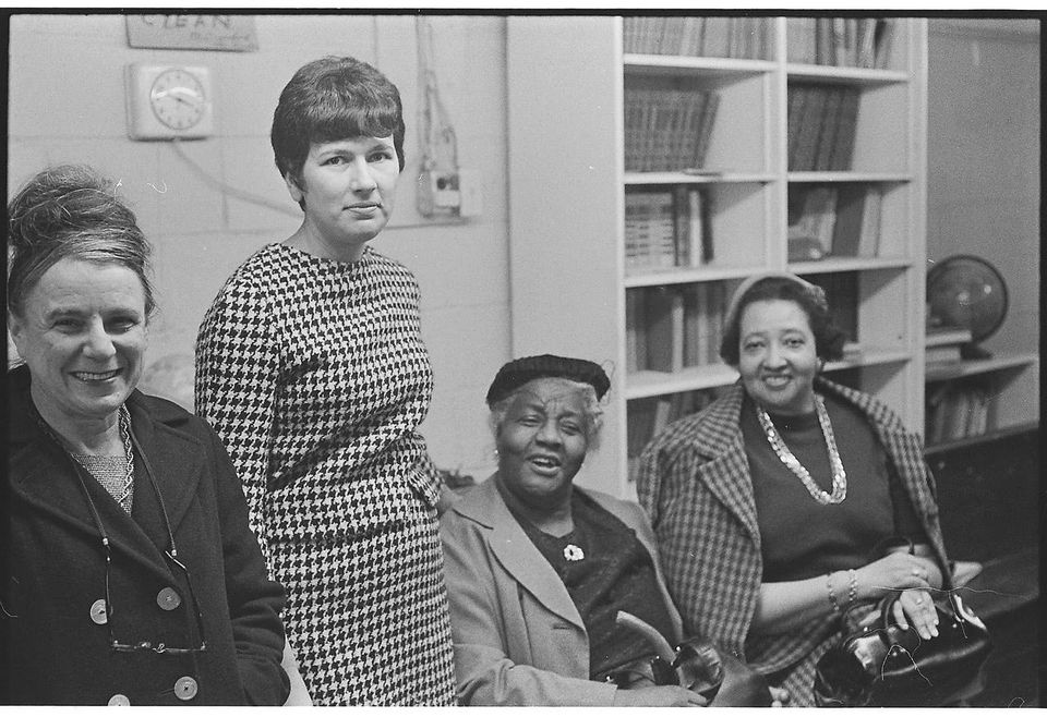 Black-and-white photograph of four women in a room with bookshelves; Joyce Bernhardt Siegel stands second from left in a houndstooth dress