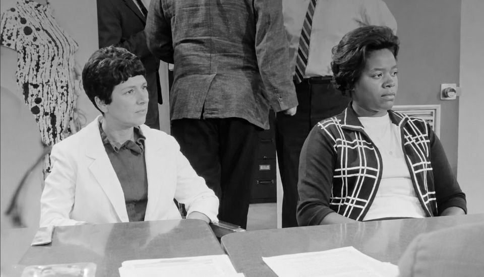 Black-and-white photograph of two women seated at a meeting table, with men standing behind them