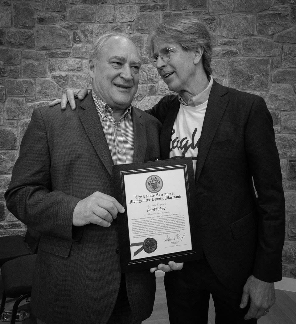 Paul Tukey, right, with his arm around another man who holds a framed certificate from the County Executive of Montgomery County, Maryland